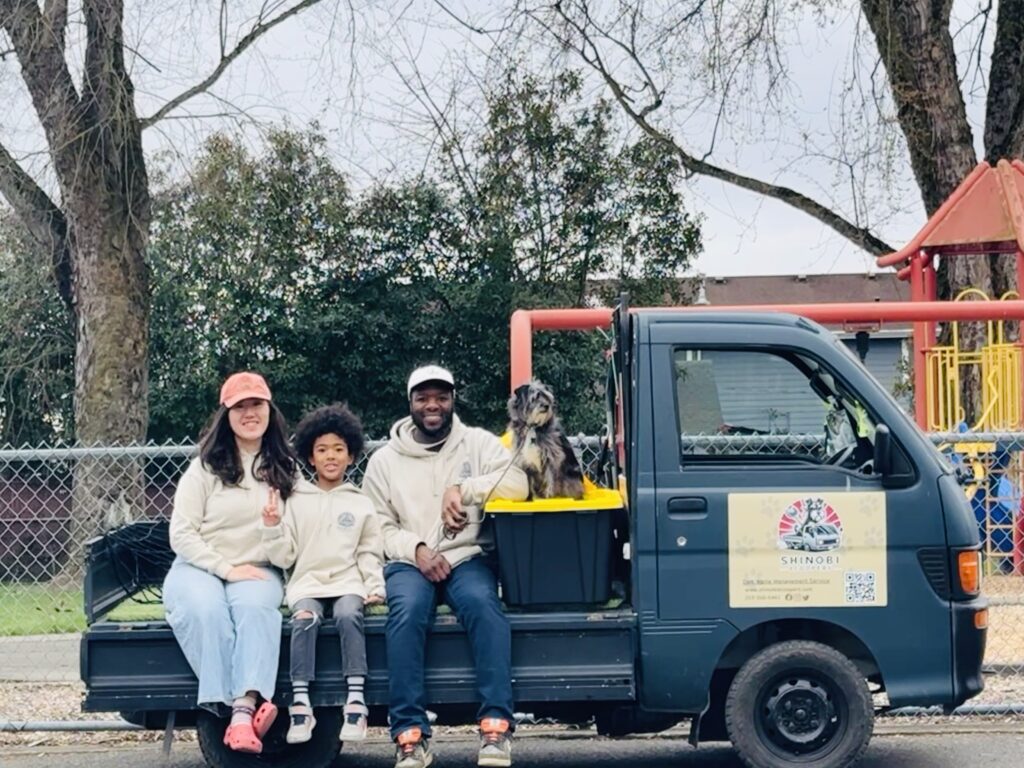 Madoka, their son Adon, Miles, and their dog Magic sitting on their 'Shinobi Scoopers' Kei Truck